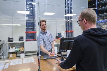 Un hombre en el mostrador de una tienda habla con un dependiente situado delante de un almacén AutoStore