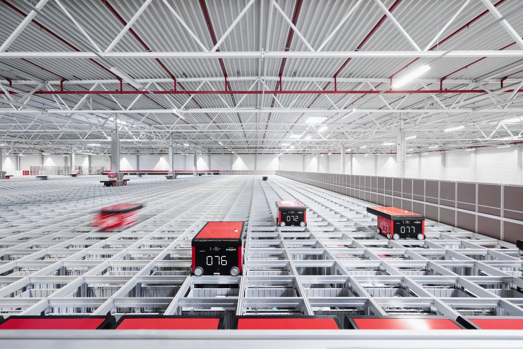 Vista general de un gran almacén automatizado con robots de AutoStore rojos en movimiento sobre una extensa rejilla metálica, bajo una estructura de techo industrial.