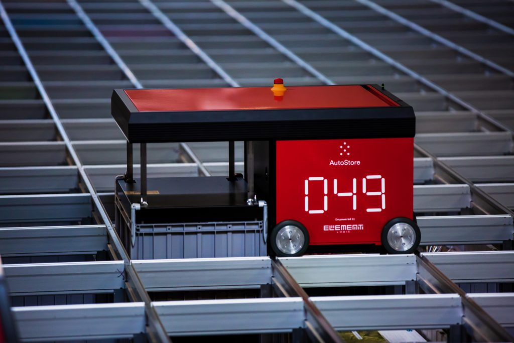 Robot de almacenamiento rojo y negro del sistema AutoStore, marcado con el número 049, desplazándose entre las estructuras metálicas grises de una rejilla de almacenamiento automatizada, con el logo de 'Element Logic' destacado en su lateral.