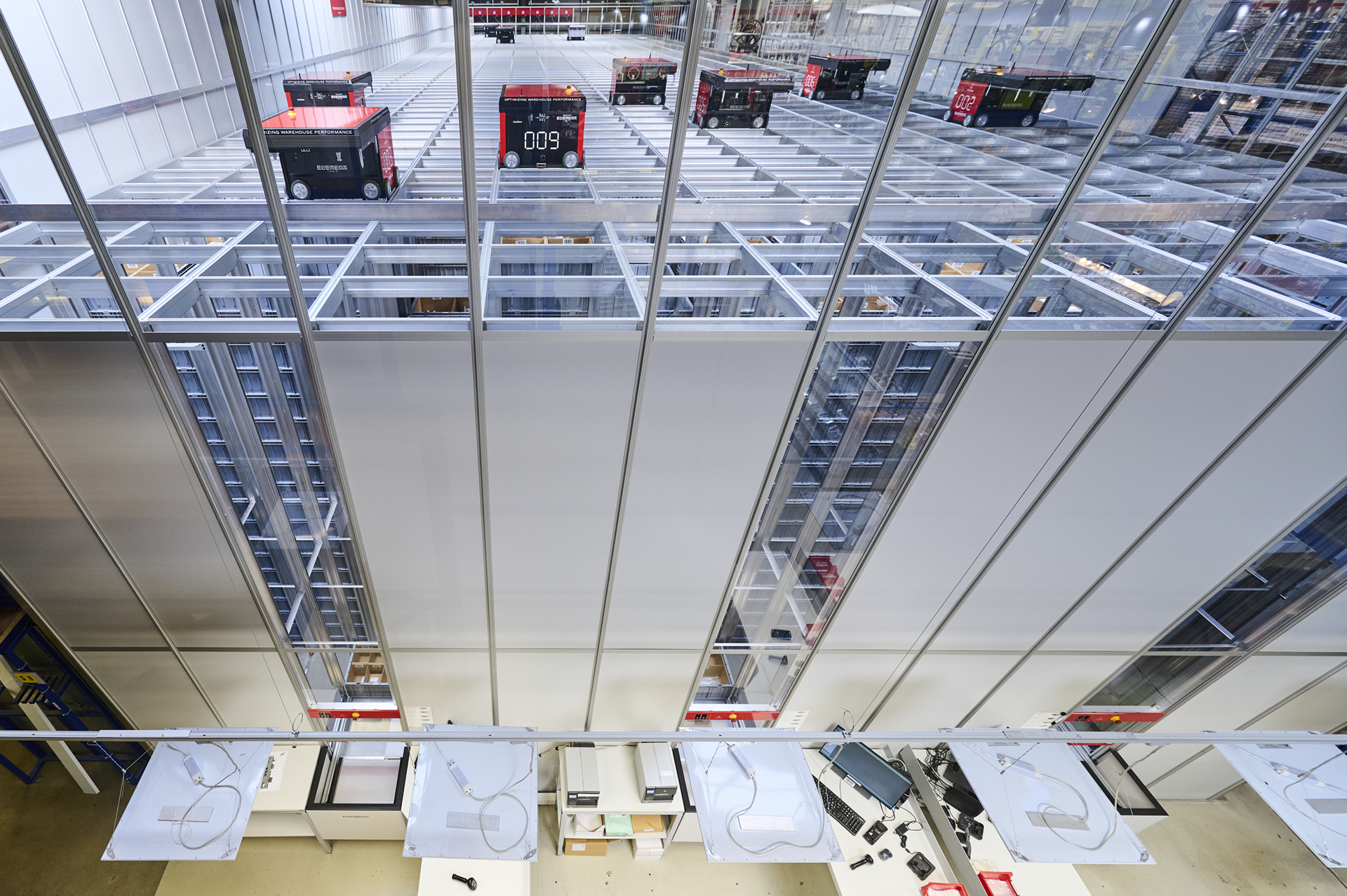 Vista de ángulo elevado de un sistema de almacenamiento automatizado de AutoStore, mostrando robots rojos desplazándose sobre una rejilla metálica con estaciones de trabajo debajo.