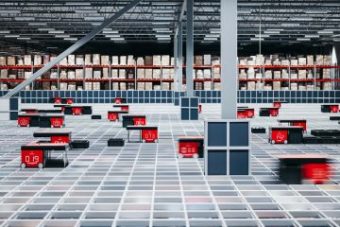"Robots de AutoStore rojos maniobrando en un sistema de almacenamiento de rejilla en un almacén con estanterías altas al fondo, mostrando una operación logística moderna y automatizada
