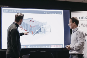 Dos hombres en una sala de conferencias, uno de ellos señalando hacia una pantalla grande que muestra el diseño en 3D de un almacén automatizado. La pantalla muestra el nombre "CBK [AutoStore]" y un número de proyecto. Hay un banner de "CBK" junto a la pantalla.